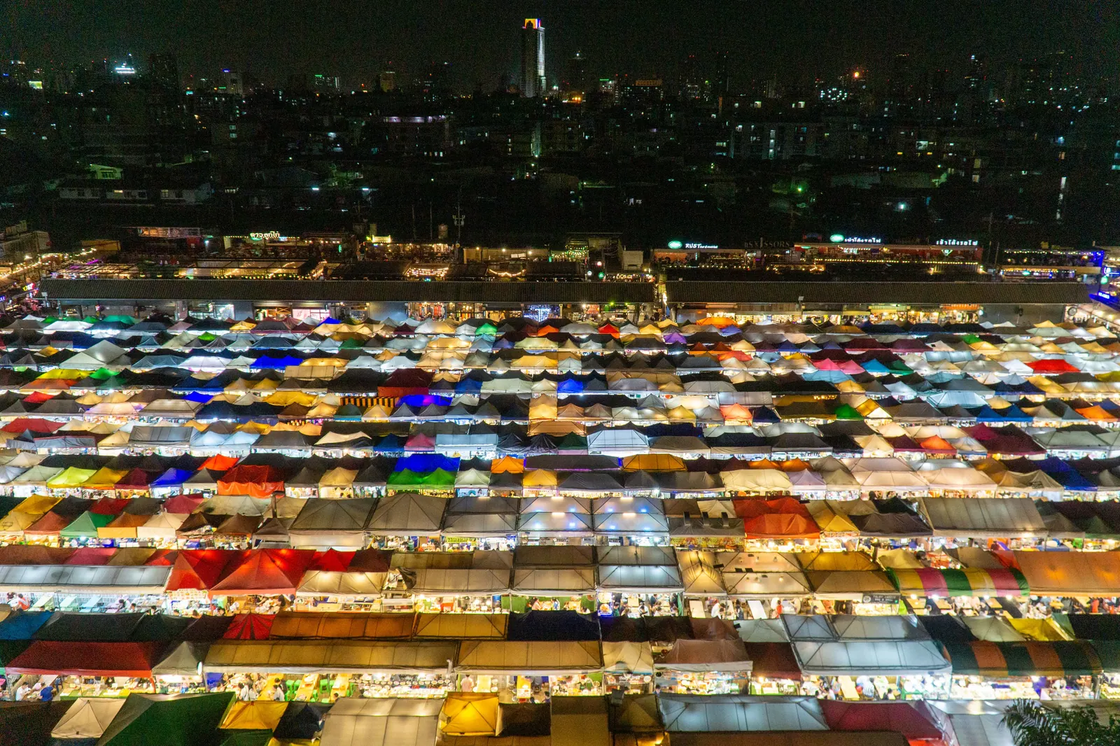 ตลาดนัดกลางคืนในไทยมองจากมุมสูง หลังคาเต็นท์เรียงเป็นแถวสว่างทุกสี มีเส้นขอบฟ้าของเมืองอยู่ไกลๆ