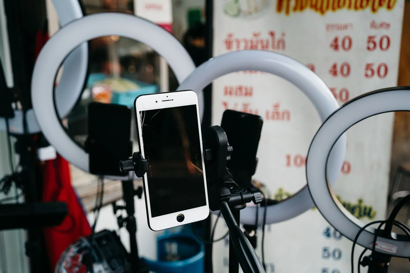 สมาร์ตโฟนบนขาตั้งหน้าไฟวงแหวนสามดวงที่ร้านข้าวมันไก่ ป้ายเมนูภาษาไทยด้านหลังเขียนราคา 40 และ 50 บาท